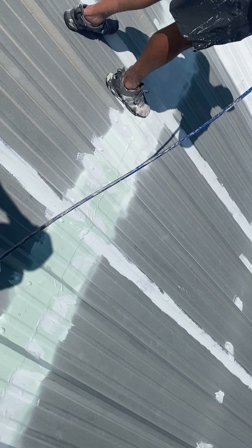 Person applying white sealant on a metal roof, showcasing shoes and gray roofing material.