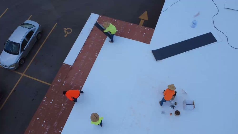 Workers installing a white roofing membrane on a commercial building from an aerial view.