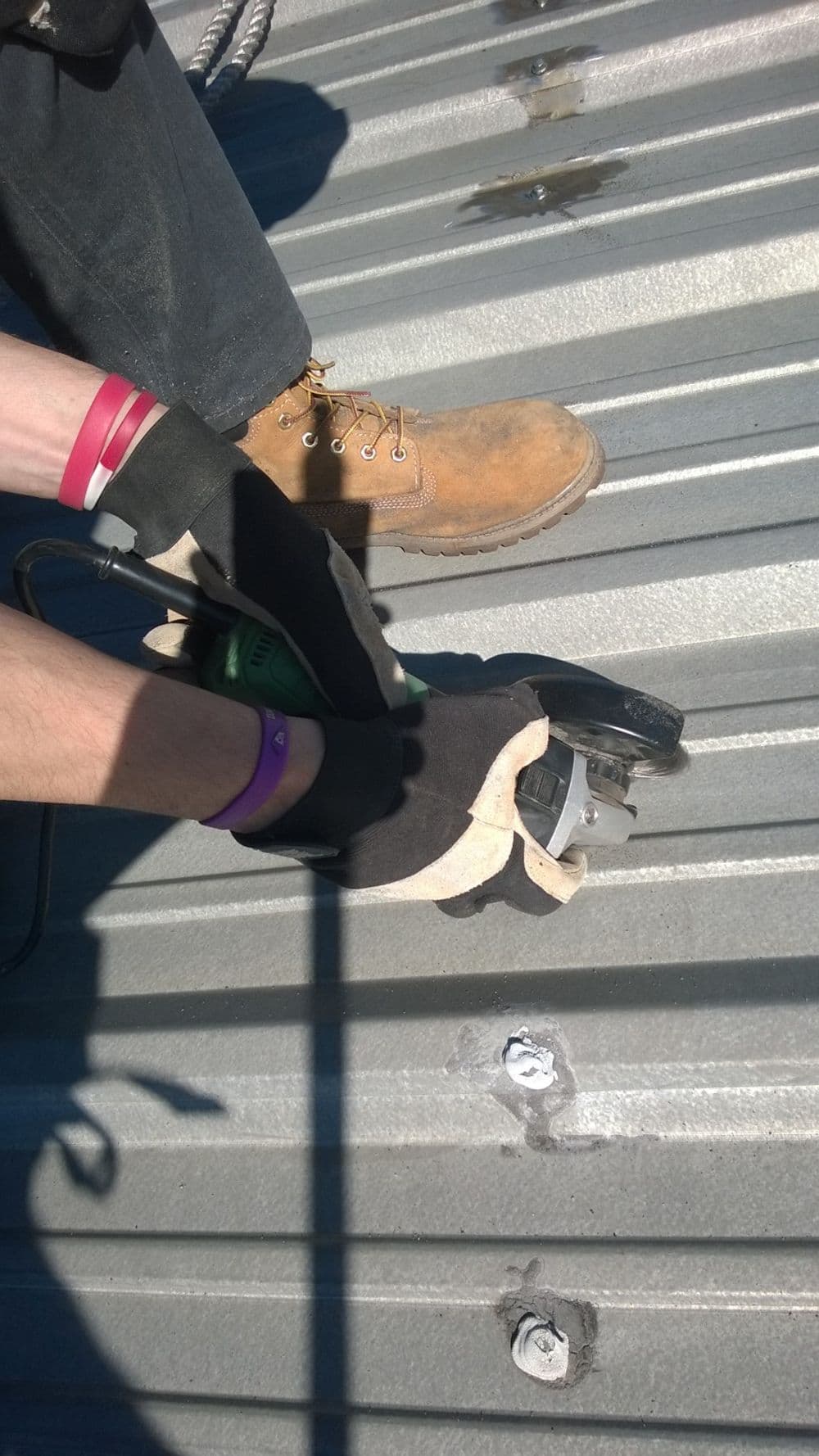 Person wearing gloves using a power tool on a metal roof, with safety boots visible.