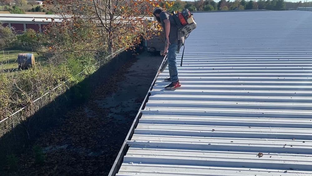 Person cleaning leaves from a flat metal roof using a leaf blower on a sunny day.