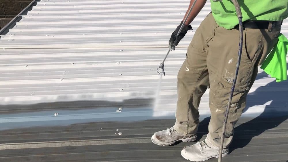 Person applying white coating to a metal roof using a spray gun for energy efficiency.