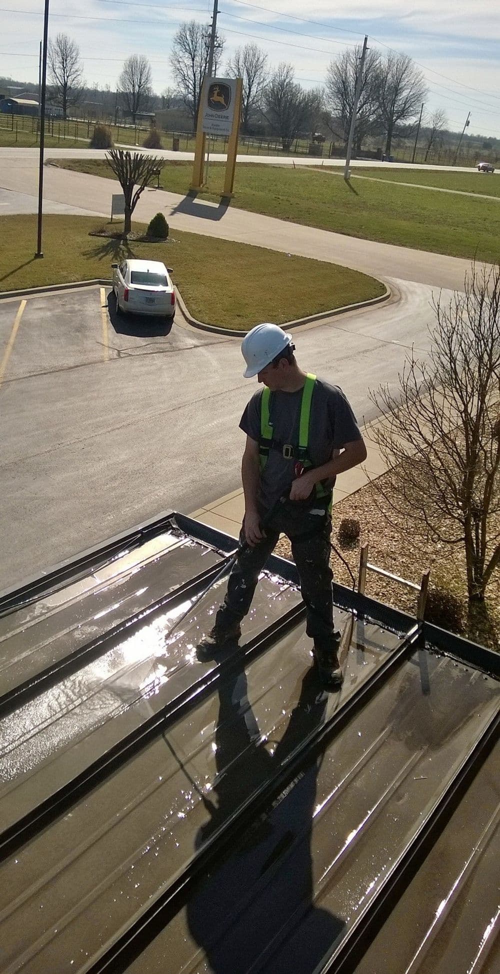 Worker in helmet and safety gear standing on a roof, showcasing a clear sky and parking lot below.