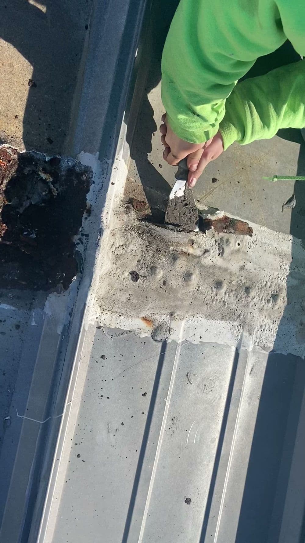 Worker repairing metal roofing with trowel, focusing on sealant application at joint.