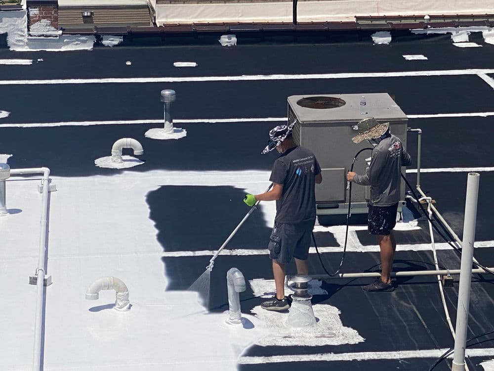 Workers applying white roof coating on a commercial building with HVAC units visible.
