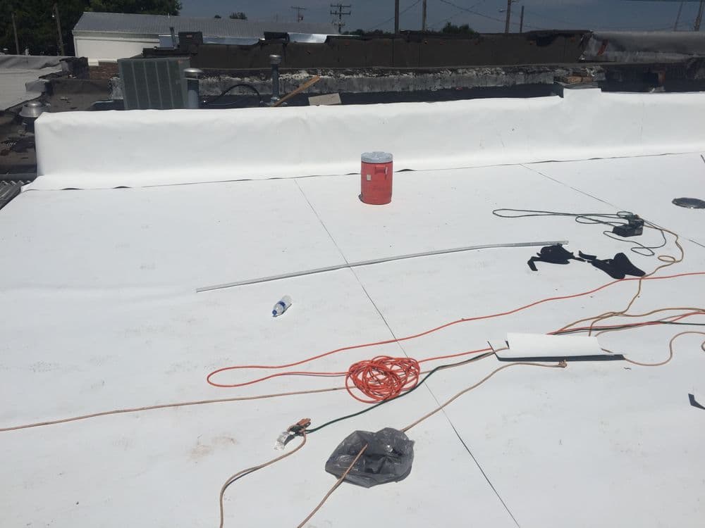 Flat roofing installation with tools, orange extension cords, and a paint bucket on a white surface.