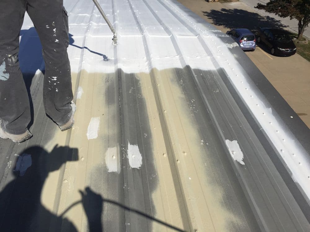 Worker spraying white paint on a metal roof, showing progress with gray and beige sections.