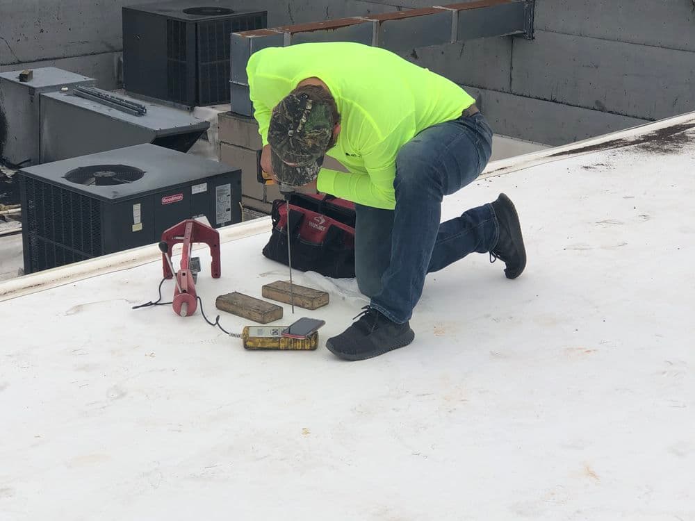 Worker in bright yellow shirt kneels on roof, using tools for maintenance near HVAC units.