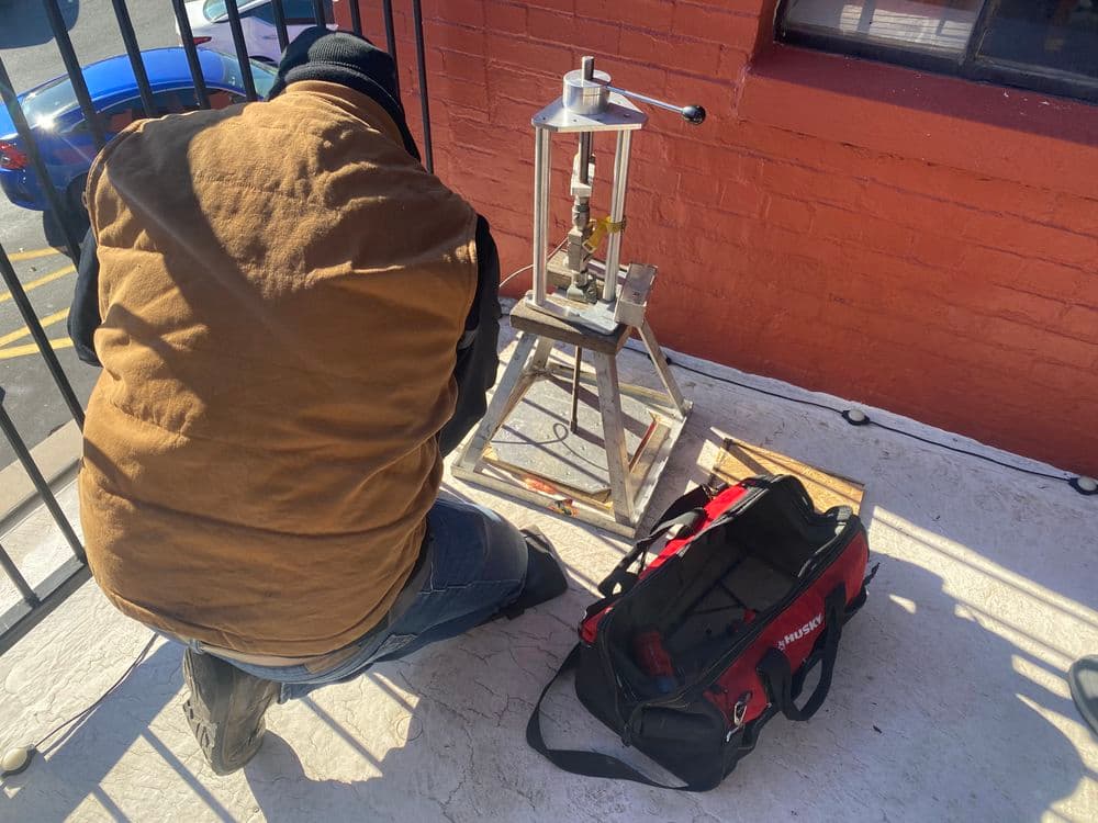 Person operating a metal press on a balcony with a tool bag nearby.