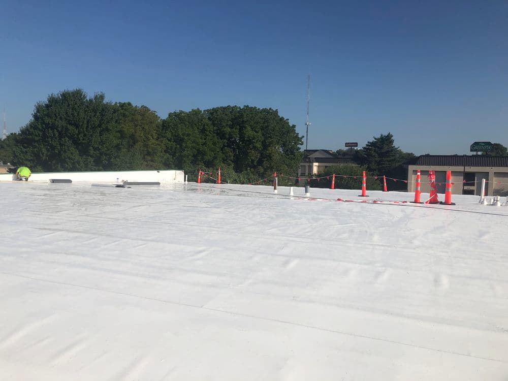 Workers on a commercial roof applying a white membrane with safety cones and clear sky.