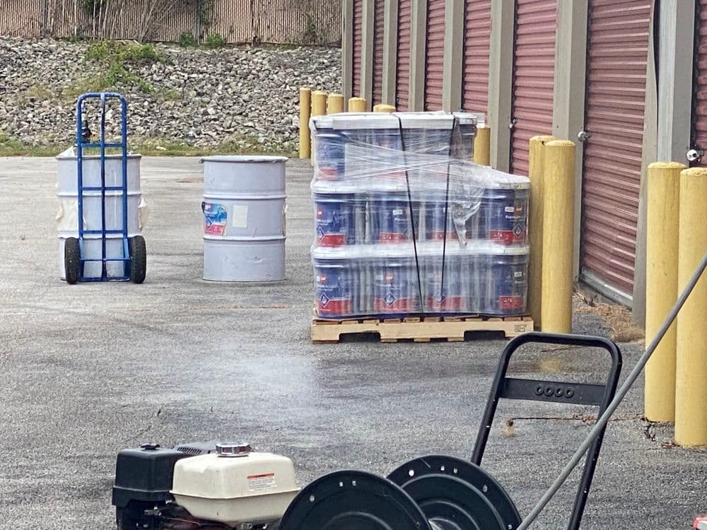 Storage area with stacked paint cans on a pallet, a drum, and a hand truck nearby.