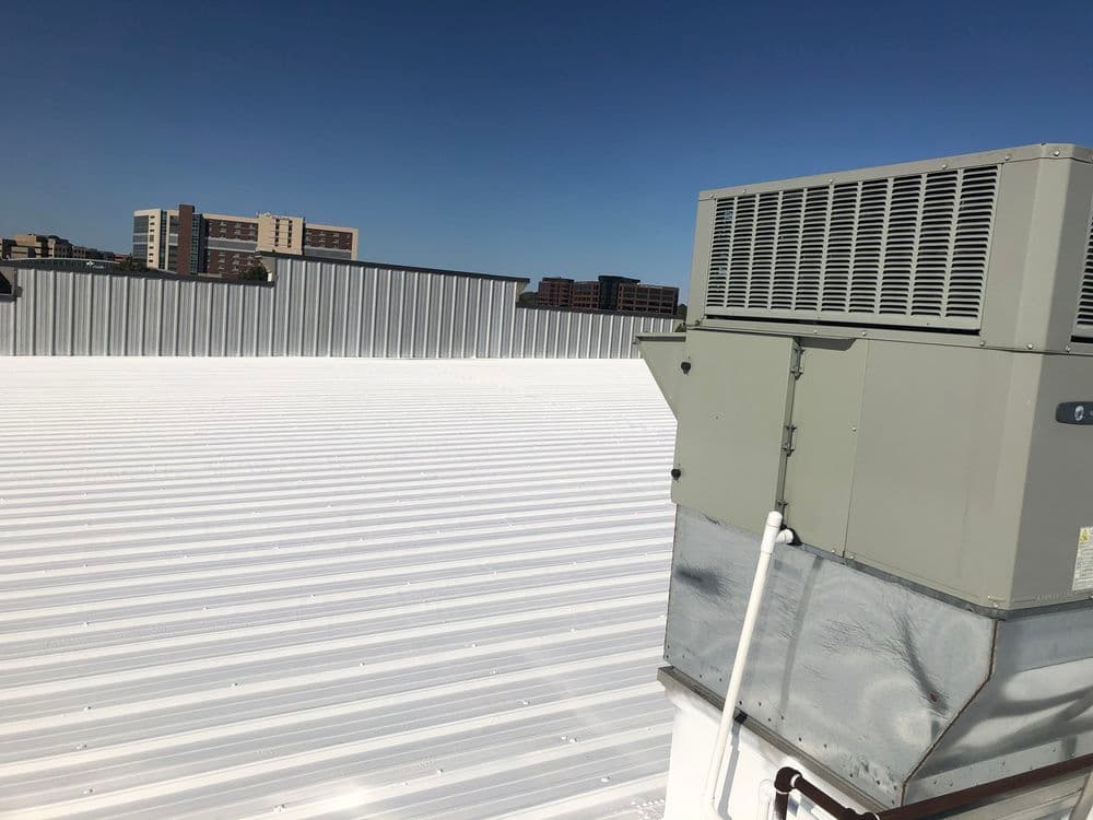 Flat white rooftop with HVAC unit against a clear blue sky and distant buildings.