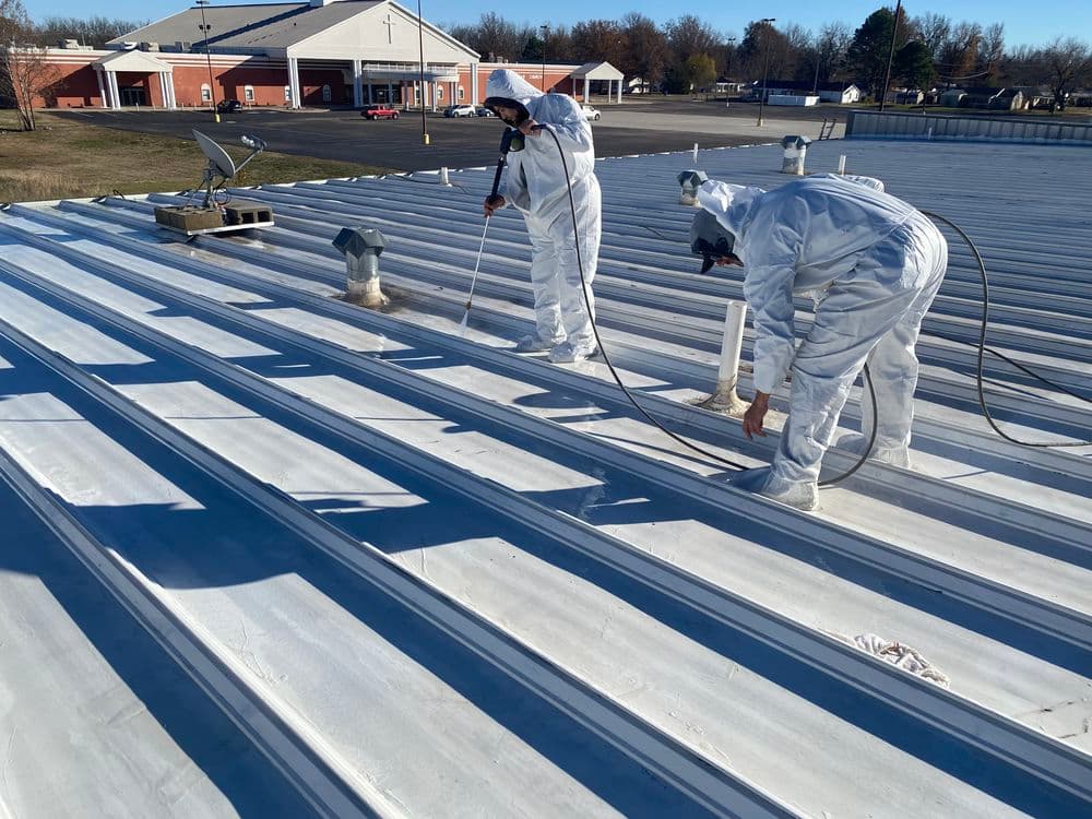 Workers in white protective suits painting a metal roof with sprayers on a sunny day.