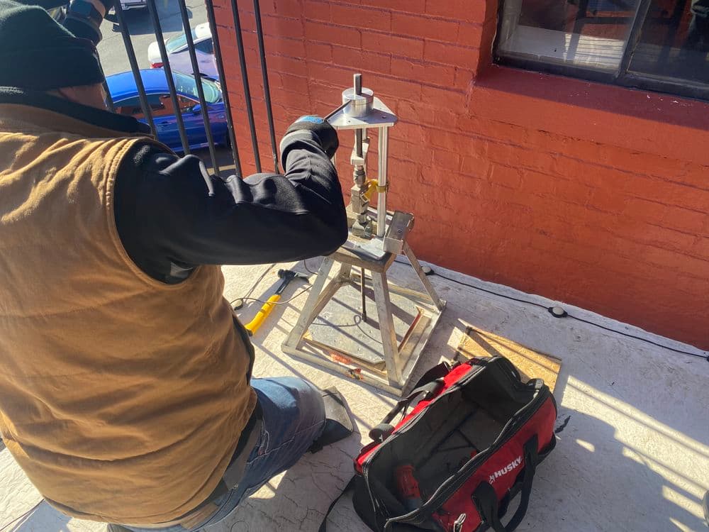Worker using a metal press machine outdoors with tools and a red bag nearby.