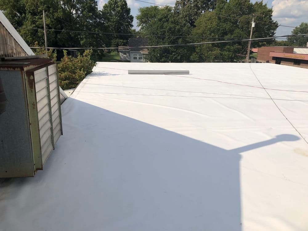 Flat white roofing surface with nearby structures and trees under a clear sky.