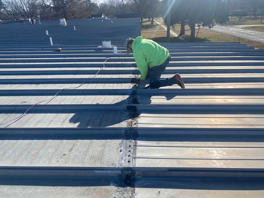 Worker in a bright green jacket inspecting seams on a metal roof under sunlight.