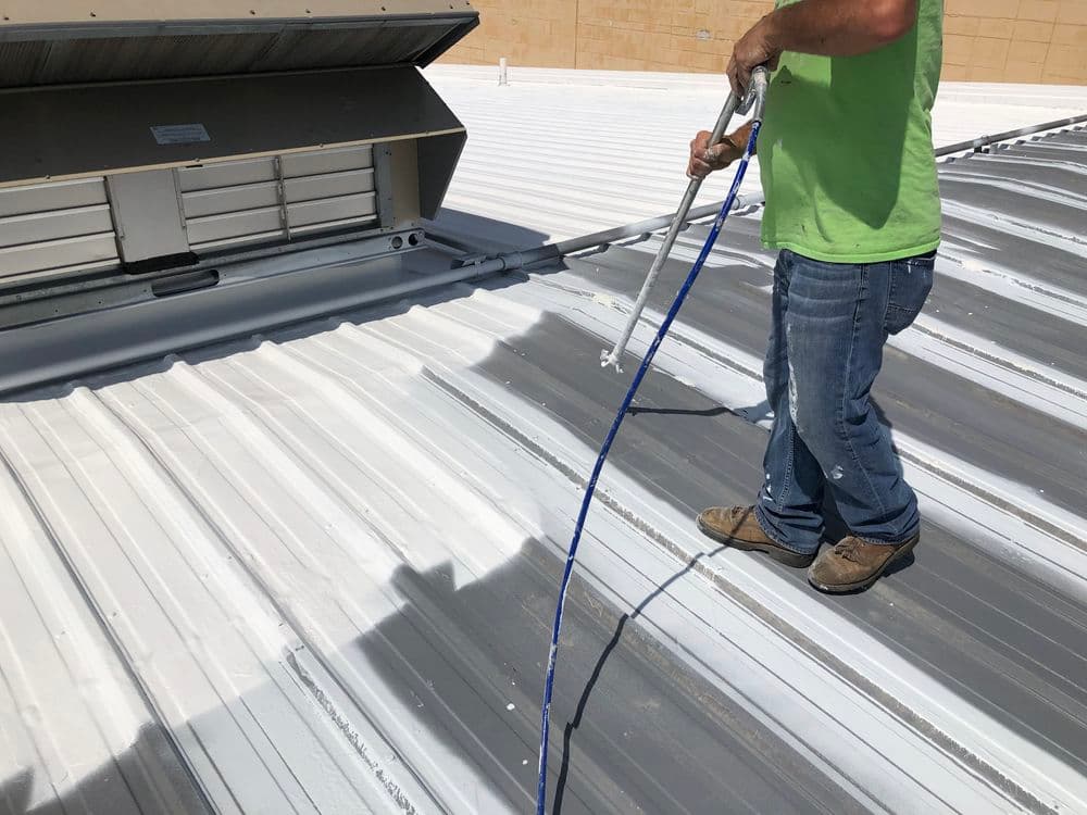 Worker applying sealant on a metal roof with spray equipment under sunlight.