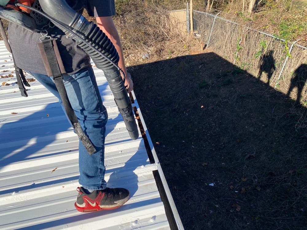 Person using a leaf blower on a metal roof, clearing debris with shadows in the background.