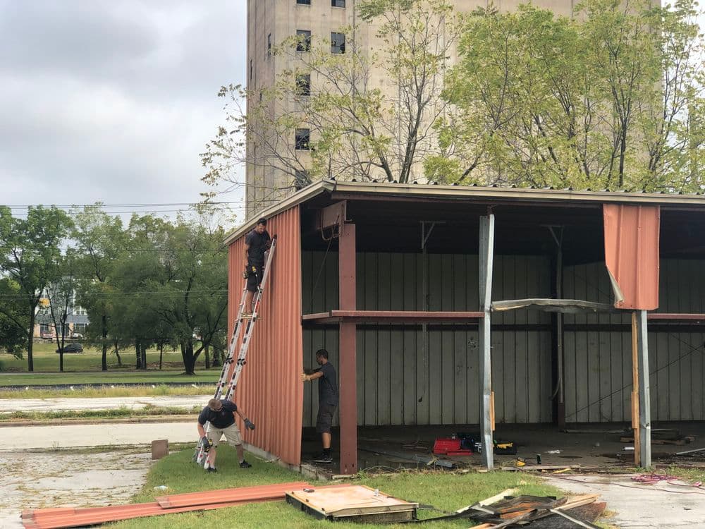 Workers renovating a rusted metal building with a ladder and tools on a cloudy day.