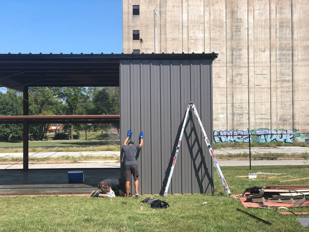 Worker installing metal siding on a building with a ladder nearby and graffiti in the background.
