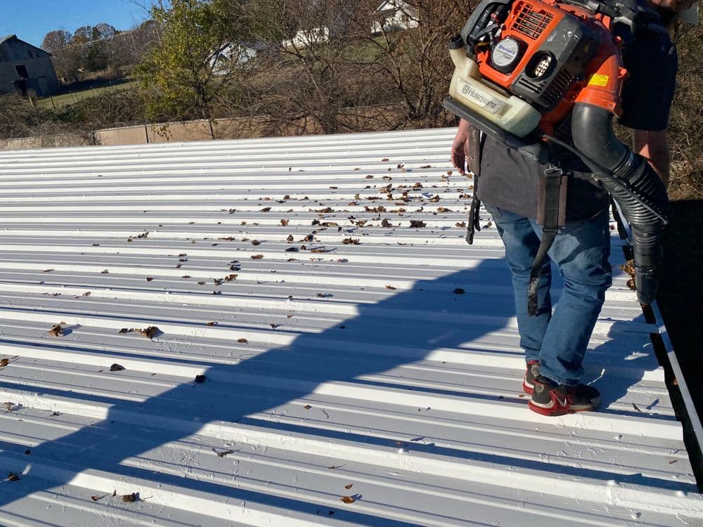 Person using a leaf blower to clean leaves off a metal roof on a sunny day.