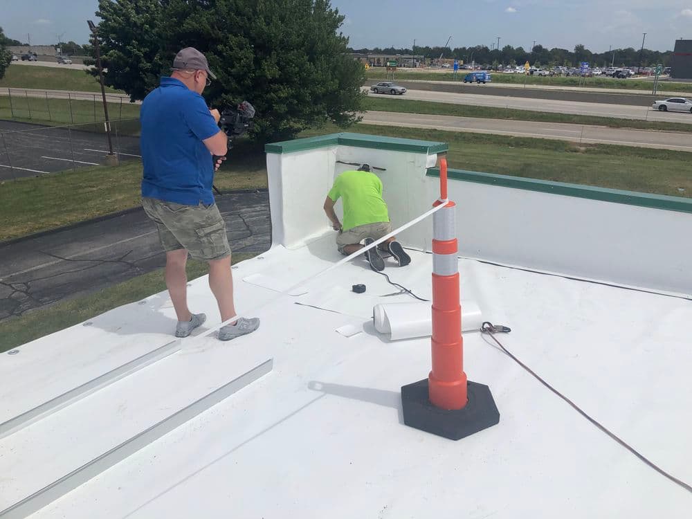 Worker repairing a roof while a cameraman films the process outdoors on a sunny day.